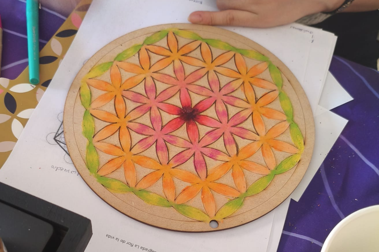 Un diseño colorido de mandala dibujado sobre una base circular de madera, con flores de tonos anaranjados, rosas y verdes, acompañado de materiales de arte en el fondo.
